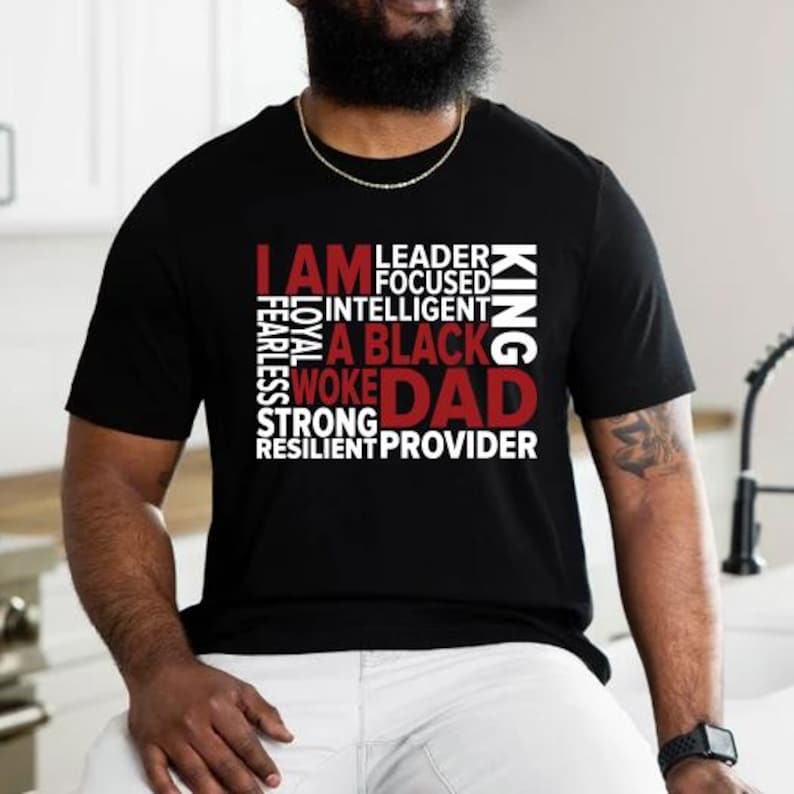 Op de afbeelding: Zwart t-shirt met de tekst "I AM A BLACK WOKE DAD" in witte en rode letters. Extra woorden zijn "Leader, Focused, Intelligent, Loyal, Fearless, Strong, Resilient, King, Provider."