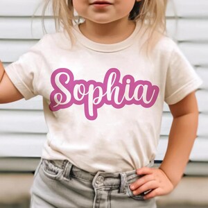 K&ouml;nnte beinhalten: Ein cremefarbenes T-Shirt mit dem Namen "Sophia" in leuchtend rosa Schrift. Der Text ist wei&szlig; umrandet und erzeugt einen starken Kontrast. Das Shirt ist kurz&auml;rmlig und scheint aus einem weichen Material zu sein.