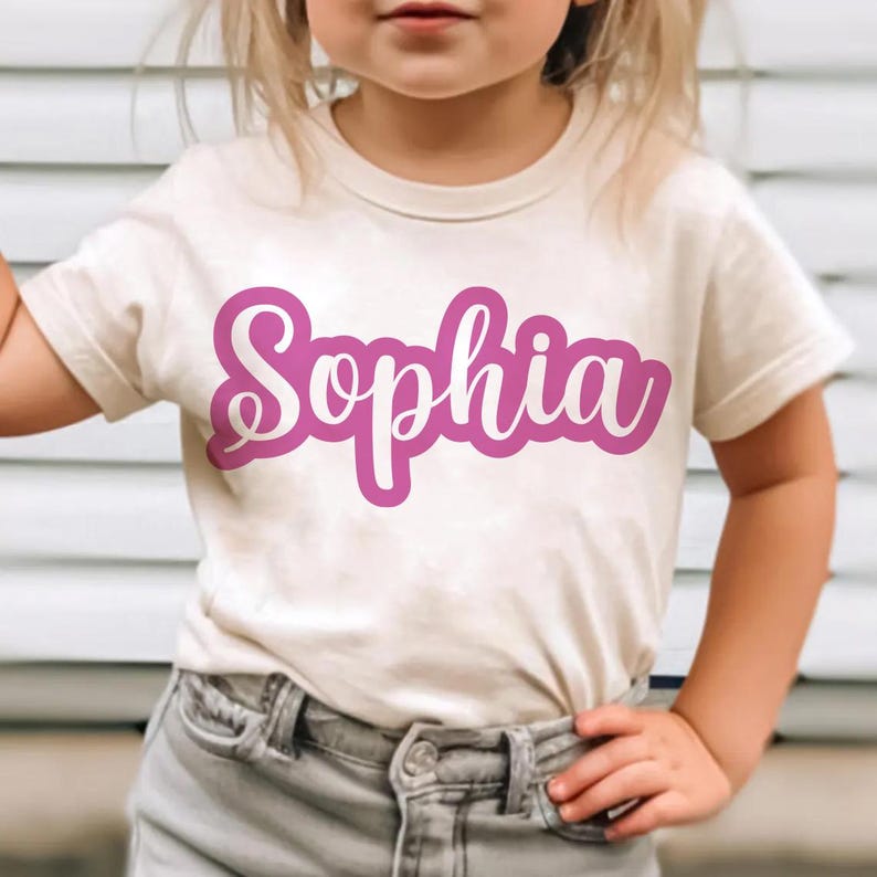 K&ouml;nnte beinhalten: Ein cremefarbenes T-Shirt mit dem Namen "Sophia" in leuchtend rosa Schrift. Der Text ist wei&szlig; umrandet und erzeugt einen starken Kontrast. Das Shirt ist kurz&auml;rmlig und scheint aus einem weichen Material zu sein.