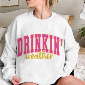 May include: White sweatshirt with the text "DRINKIN' weather" in pink and gold letters.