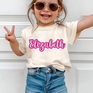 K&ouml;nnte beinhalten: Ein cremefarbenes T-Shirt mit dem Namen "Elizabeth" in leuchtend rosa Schrift. Das Shirt wird mit blauen Jeans kombiniert. Die Person auf dem Bild tr&auml;gt eine Sonnenbrille und macht ein Friedenszeichen mit ihren Fingern.