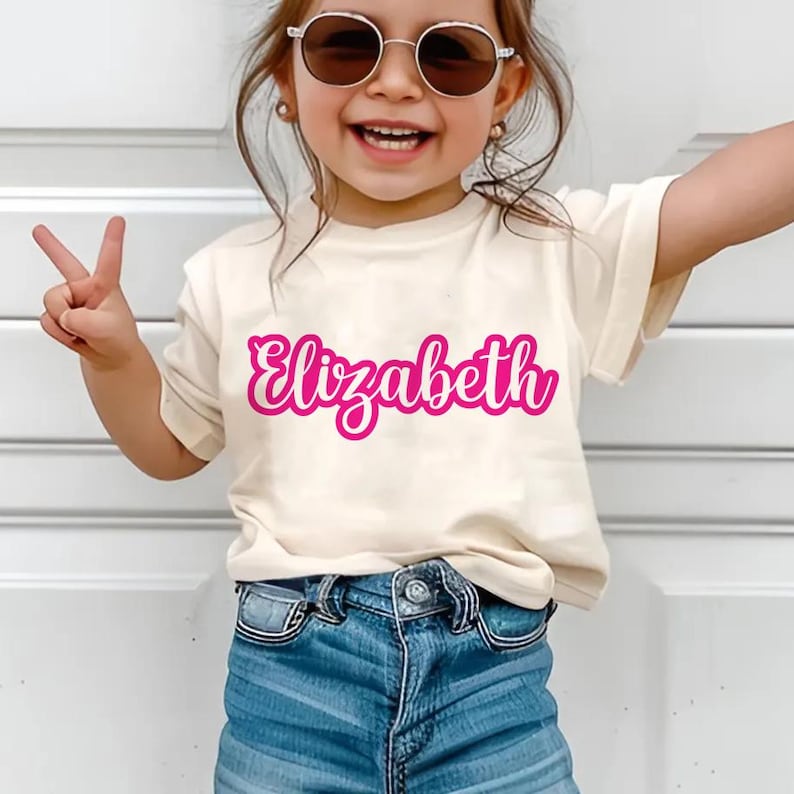 K&ouml;nnte beinhalten: Ein cremefarbenes T-Shirt mit dem Namen "Elizabeth" in leuchtend rosa Schrift. Das Shirt wird mit blauen Jeans kombiniert. Die Person auf dem Bild tr&auml;gt eine Sonnenbrille und macht ein Friedenszeichen mit ihren Fingern.