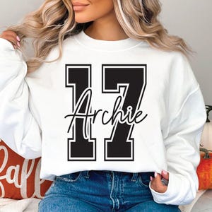 May include: White sweatshirt with the number 17 in black block letters with a white outline. The name "Archie" is written in cursive script across the center of the number. The sweatshirt is a casual style.