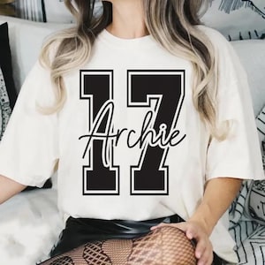 May include: Cream-colored t-shirt featuring the number "17" in a bold, black, block-letter style with a white outline. The name "Archie" is scripted in black cursive across the numbers. The shirt has short sleeves and a relaxed fit.