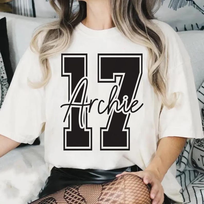 May include: Cream-colored t-shirt featuring the number "17" in a bold, black, block-letter style with a white outline. The name "Archie" is scripted in black cursive across the numbers. The shirt has short sleeves and a relaxed fit.
