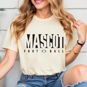 May include: Cream-colored t-shirt with the word "MASCOT" in bold, black capital letters, above the words "FOOTBALL" and a football graphic. The shirt is a casual style.