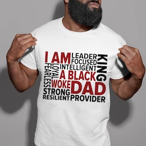 Op de afbeelding: Wit T-shirt met de rode woorden "I AM", gevolgd door een lijst met positieve eigenschappen in zwart, en de rode woorden "A BLACK DAD". Het woord "KING" is verticaal in zwart gedrukt.