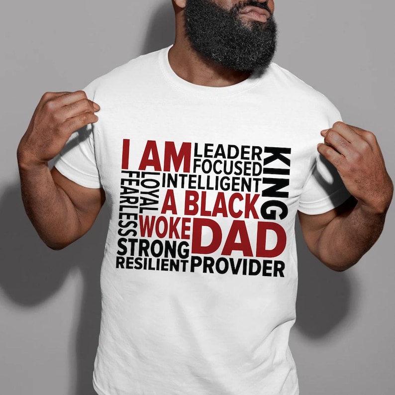 Op de afbeelding: Wit T-shirt met de rode woorden "I AM", gevolgd door een lijst met positieve eigenschappen in zwart, en de rode woorden "A BLACK DAD". Het woord "KING" is verticaal in zwart gedrukt.
