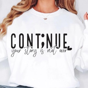 以下が含まれることがあります： 「CONTINUE your story is not over」という黒い文字が書かれた白いスウェットシャツ。CONTINUEのIの部分はセミコロンに置き換えられ、overの後に小さなハートマークが付けられています。