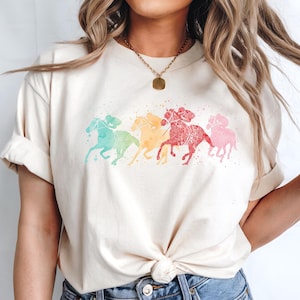 May include: A white t-shirt with a watercolor design of five horses running in a line. The horses are different colors, including red, orange, yellow, green, and blue.