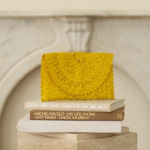 May include: A bright yellow clutch purse made of woven fabric with a slightly rounded shape. The purse is sitting on top of a stack of books.