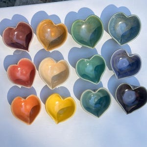 May include: A collection of twelve heart-shaped ceramic bowls in various colors, including red, orange, yellow, green, and blue. The bowls are arranged on a white surface, with shadows cast from above. Each bowl has a glossy finish.