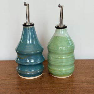 May include: Two ceramic oil bottles, one teal and one green, with stainless steel pour spouts. The teal bottle has a stacked design, while the green bottle has horizontal ridges. Both bottles sit on a wooden surface.