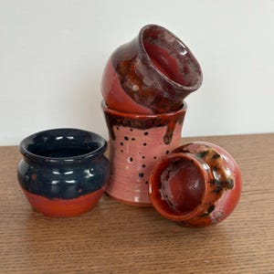May include: Four small ceramic cups in various shades of red, pink, and black. The cups are stacked on top of each other, with the top cup tilted to the side.