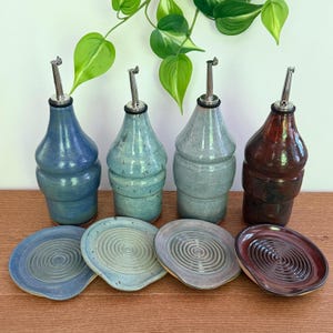 May include: Four ceramic oil dispensers with stainless steel pour spouts, each paired with a matching circular spoon rest featuring a concentric spiral pattern. The set includes one blue, one light green, one grey, and one dark reddish-brown piece.