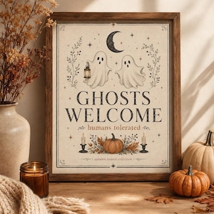 Può includere: Una stampa artistica incorniciata con due fantasmi, una luna crescente, stelle e una zucca. Il testo recita GHOSTS WELCOME, humans tolerated e autumn haunts collection. Il design include dettagli botanici e due candele accese.