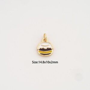 Puede incluir: Charm dorado en forma de postre en capas, con detalles de esmalte blanco, marrón y amarillo. El charm tiene un lazo para sujetarlo a una joya. El texto "Size: 14.8x10x2mm" es visible.