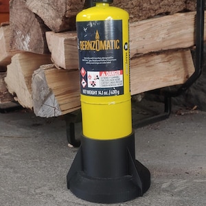 May include: A yellow Bernzomatic TS4000 propane torch with a black handle and a red on/off switch. The torch is sitting on a black base and is next to a pile of firewood.