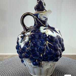 Op de afbeelding: Een blauwe en witte keramische karaf met gouden rand en een druivenontwerp. De karaf heeft een deksel en een handvat. De karaf staat op een grijze en witte ronde placemat.