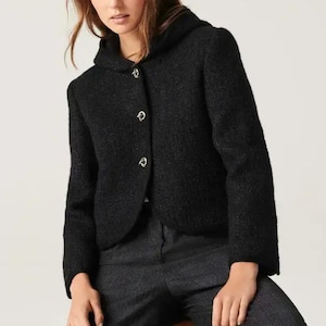 Op de afbeelding: Zwarte jas met capuchon en drie decoratieve knopen. De jas is gemaakt van een textuurstof en wordt gecombineerd met een donkergrijze broek. Het model zit op een houten kruk.