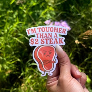 May include: A white sticker with the text "I'M TOUGHER THAN A $2 STEAK" and a cartoon steak illustration. The steak is red with a brown outline, arms crossed, and an angry expression. The sticker is held against a background of green foliage.