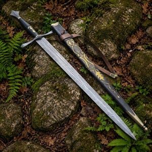 May include: A longsword with a patterned Damascus steel blade, a black grip, and a crossguard. The matching black scabbard features gold-colored metal accents, a decorative vine-like design, and a brown leather strap for carrying the weapon.