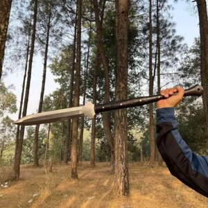 May include: A large, silver spear with a broad blade and a dark, textured handle. The spear is held up against a backdrop of tall trees in a forest setting. The handle appears to be wrapped in a dark material.