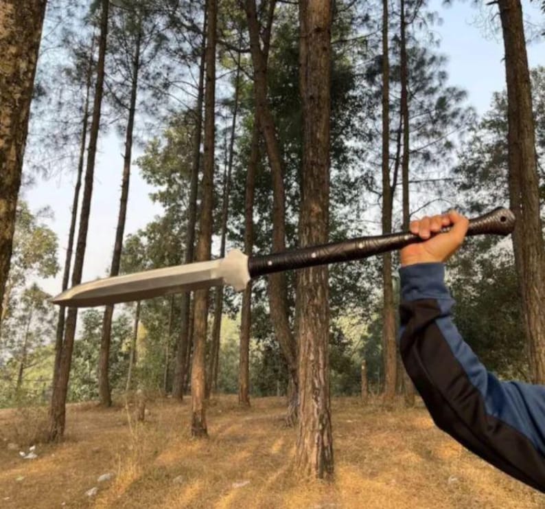 May include: A large, silver spear with a broad blade and a dark, textured handle. The spear is held up against a backdrop of tall trees in a forest setting. The handle appears to be wrapped in a dark material.