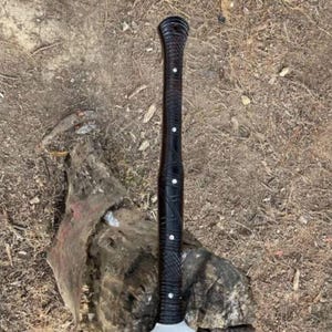 May include: A black axe with a dark wooden handle featuring white accents. The handle has a textured design. The axe head is silver. The axe is resting on a piece of wood.