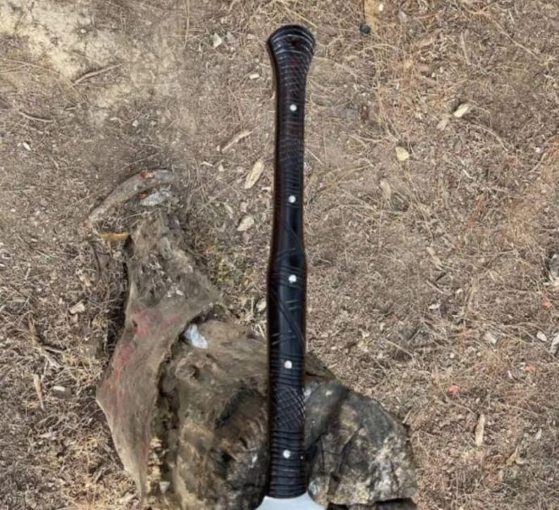 May include: A black axe with a dark wooden handle featuring white accents. The handle has a textured design. The axe head is silver. The axe is resting on a piece of wood.