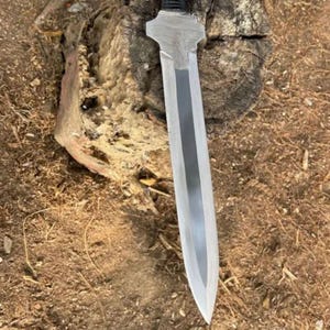May include: A silver sword with a broad blade and a black handle. The sword is positioned on a tree stump, with brown mulch surrounding it.