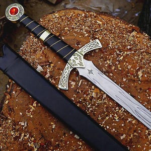 May include: A decorative sword with a silver blade and ornate gold and silver hilt. The handle is wrapped in black leather with gold accents. The sword rests on a wooden surface with a black sheath.