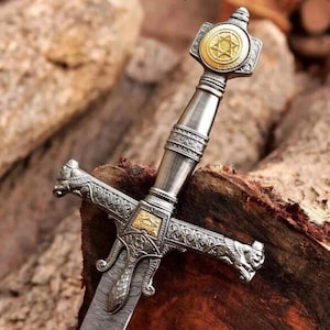 Puede incluir: Primer plano detallado de una espada decorativa con empuñadura y guarda de plata y oro. La empuñadura presenta un emblema dorado con un diseño de estrella. La guarda tiene tallas ornamentadas y detalles de cabeza de león. La hoja tiene un diseño.