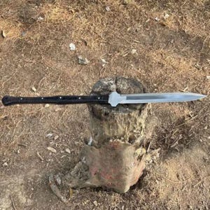 May include: A long knife with a silver blade and a black handle. The knife is placed on a weathered wooden stump. The handle has a textured pattern and is secured with small silver rivets. The blade is sharp and pointed.