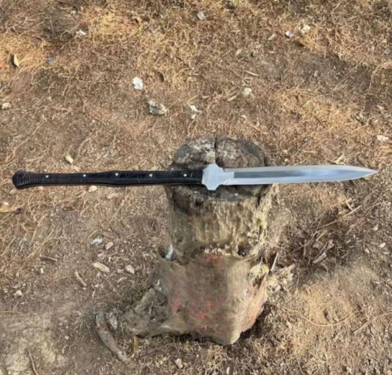 May include: A long knife with a silver blade and a black handle. The knife is placed on a weathered wooden stump. The handle has a textured pattern and is secured with small silver rivets. The blade is sharp and pointed.