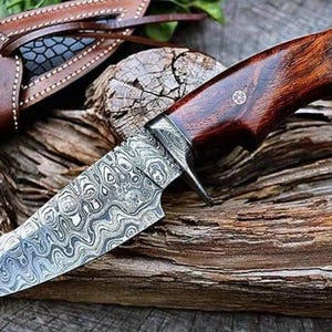 May include: A Damascus steel knife with a dark wood handle and leather sheath. The blade has a distinctive swirling pattern. The knife is displayed on weathered wood, highlighting its rustic charm. The handle is dark brown.