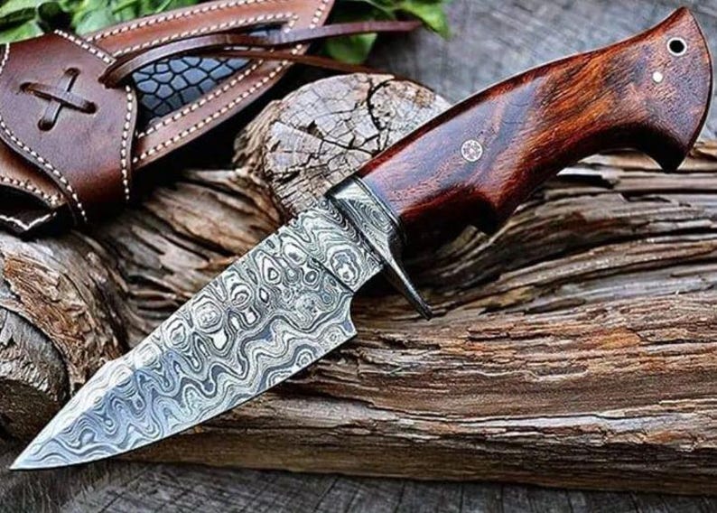 May include: A Damascus steel knife with a dark wood handle and leather sheath. The blade has a distinctive swirling pattern. The knife is displayed on weathered wood, highlighting its rustic charm. The handle is dark brown.
