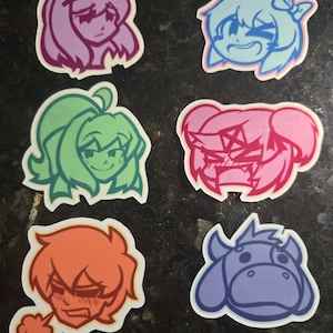 Puede incluir: Seis pegatinas de personajes de dibujos animados de colores con diferentes expresiones. Las pegatinas están sobre una superficie gris oscuro. Los personajes incluyen una niña con cabello rosa, una niña con cabello azul, una niña con cabello verde, una niña con cabello rosa, un niño con cabello naranja y una vaca azul.
