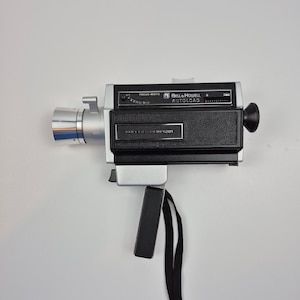 Puede incluir: Una cámara de cine vintage Bell & Howell Autoload Focus-Matic. La cámara es plateada y negra con un asa y correa negras. La cámara tiene el texto "Bell & Howell Autoload" y "Focus-Matic" en la parte superior.