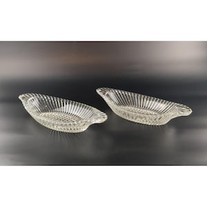 May include: Two clear glass oval serving dishes. Each dish has a textured, ribbed design on the sides and a patterned base. The dishes have small handles on each end. The background is dark.