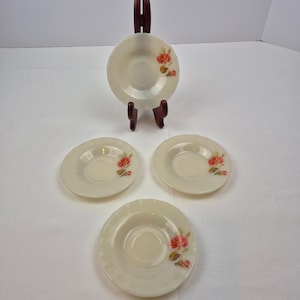 May include: Four off-white saucers with a floral rose design. One saucer is displayed on a dark brown stand. The roses are red and pink with green leaves. The saucers have a scalloped edge.