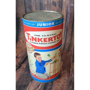 Könnte beinhalten: Eine Vintage TinkerToy Junior Builder Set Blechdose. Der zylindrische Behälter hat ein rot-weißes Design mit den Worten "TinkerToy" und "The Classic Construction Set". Ein kleiner Junge wird beim Spielen mit dem Spielzeug gezeigt. Die Dose steht auf einer Holzoberfläche.