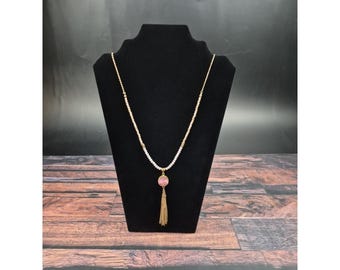 Puede incluir: Un collar dorado con una cadena de cuentas blancas y un colgante de piedra rosa. El colgante está adornado con una borla dorada. El collar se exhibe en un expositor de joyas negro sobre un fondo oscuro.