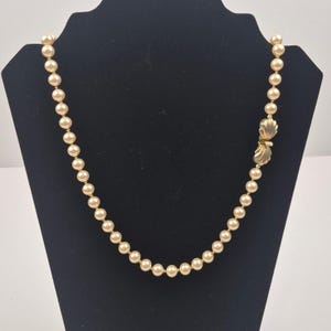 Puede incluir: Un collar de perlas color crema con un cierre dorado en forma de concha. El collar se exhibe en un maniquí negro. Las perlas están espaciadas uniformemente y tienen un tamaño constante. El cierre presenta un diseño de concha.