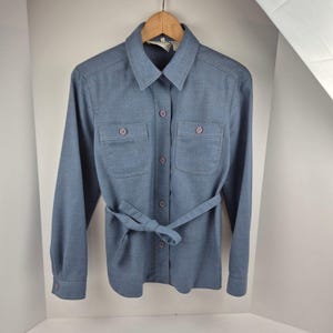 Puede incluir: Camisa azul claro con mangas largas, dos bolsillos en el pecho y cinturón a juego. La camisa está colgada de una percha de madera. Los botones y los detalles de los bolsillos son de un azul más oscuro.