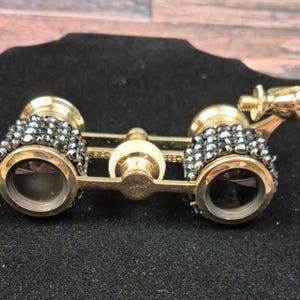 Puede incluir: Unos prismáticos dorados con detalles de cristales negros alrededor de las lentes. Los prismáticos de estilo vintage tienen una rueda de enfoque central y un pequeño botón de ajuste. El artículo está sobre un fondo negro.