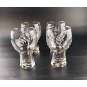 May include: Four clear glass stemware glasses with a rounded bowl and a short stem. Each glass has a large, stylized letter "S" etched on the bowl. The glasses are arranged on a dark surface, with the light reflecting off the glass.