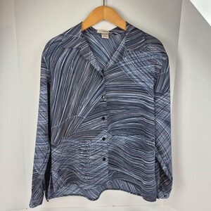 May include: A button-down shirt with a blue and white abstract pattern. The long-sleeved shirt is hanging on a wooden hanger. The shirt has a collared neckline and black buttons.