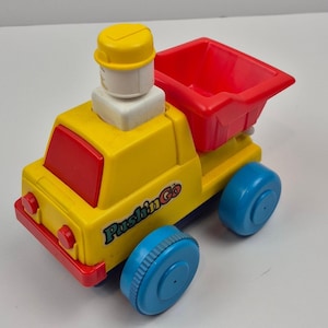May include: A yellow and red toy dump truck with blue wheels. The truck has a red dump bed and cab, with the words "Pushin' Go" printed on the side. A yellow and white structure sits on top of the cab.