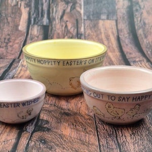 Puede incluir: Tres cuencos de cerámica con diseños de Pascua. El cuenco más grande es amarillo con el texto "Hoppity Easter's on it's way". El cuenco mediano es rosa con el texto "Peeping out to say Happy Easter". El cuenco más pequeño es rosa con el texto "Happy Easter Wishes".